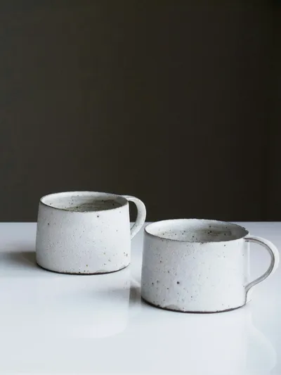 Two squat white speckled stoneware mugs with loop handles and dark clay rims on a white surface against a dark wall