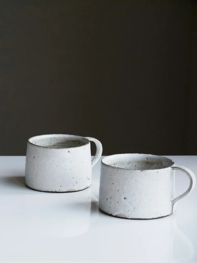 Two squat white speckled stoneware mugs with loop handles and dark clay rims on a white surface against a dark wall