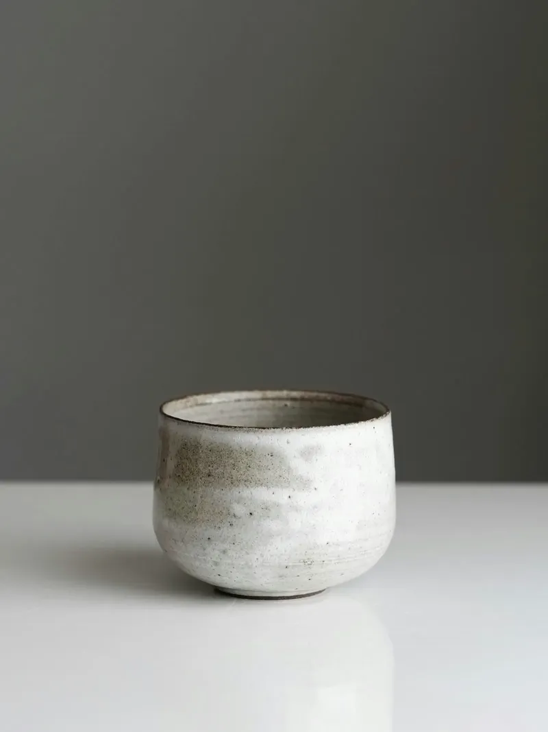Small handleless tea bowl in speckled white glaze over grey stoneware, with a dark rim, on a white surface