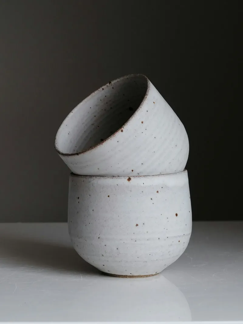 Two speckled white ceramic cups stacked, the top one tilted, with brown-edged rims on a white surface against a dark wall