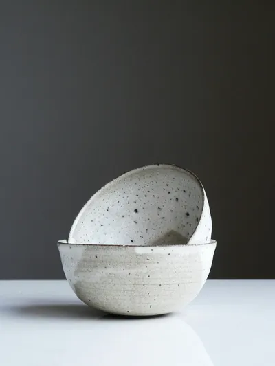 Two speckled white ceramic bowls, one tilted and resting inside the other, against a dark grey wall on a white surface