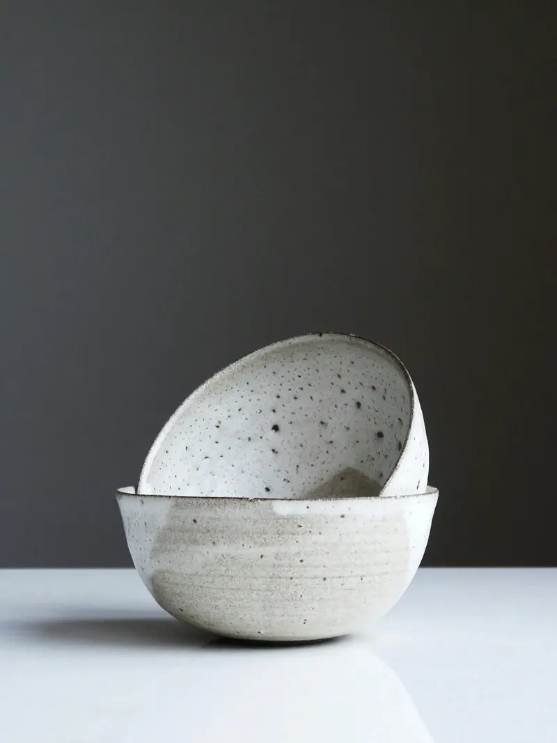 Two speckled white ceramic bowls, one tilted and resting inside the other, against a dark grey wall on a white surface