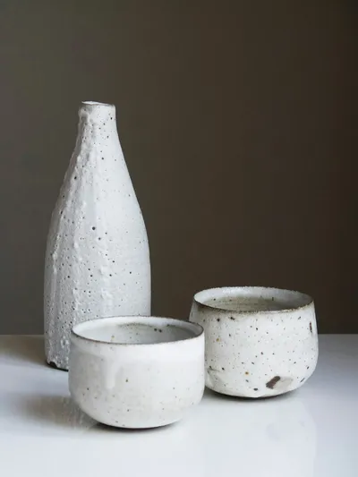 Tall white speckled bottle with a beaded crawled glaze beside two small speckled white cups on a white surface
