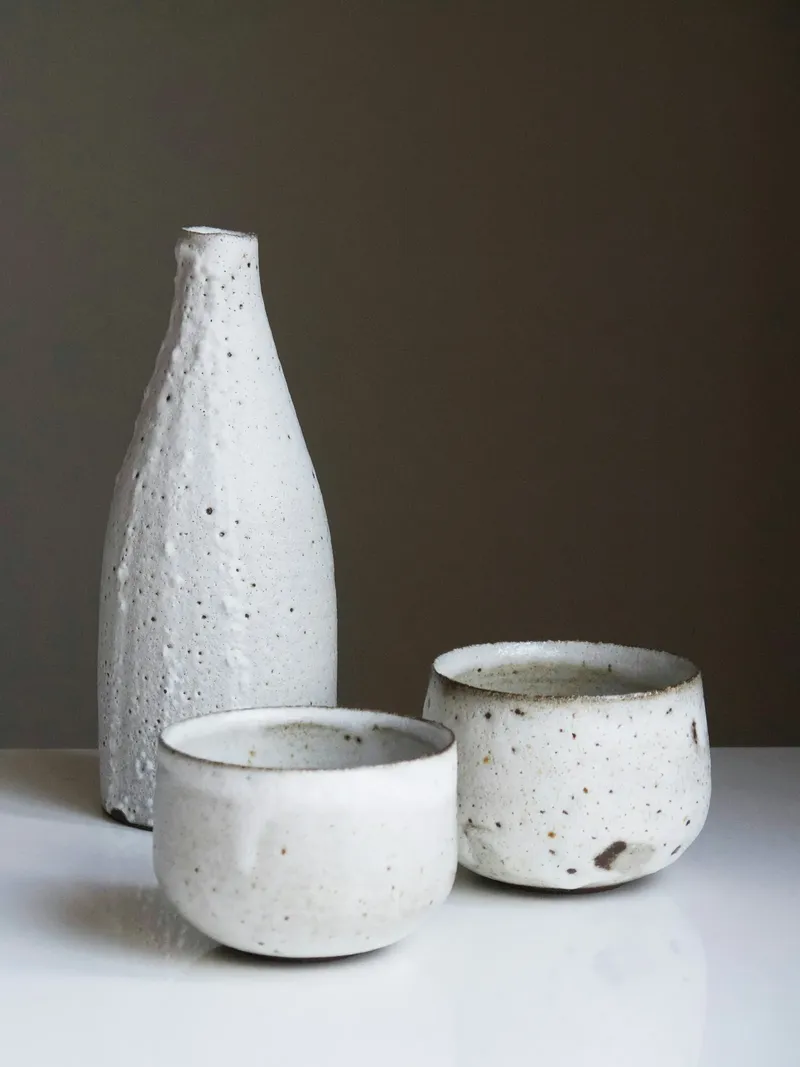 Tall white speckled bottle with a beaded crawled glaze beside two small speckled white cups on a white surface