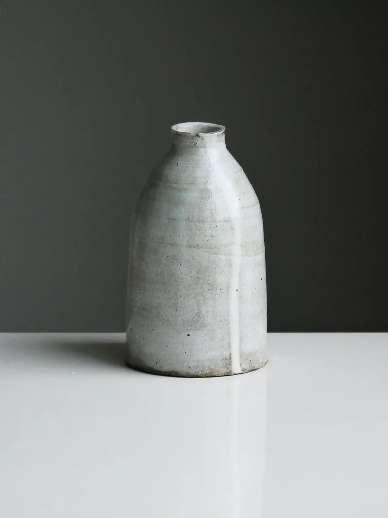 Grey-white bottle vase with a narrow neck, speckled satin glaze and one drip, on a white surface against dark grey