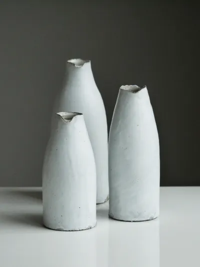 Three matte white bud vases of different heights with pinched, uneven lips, on a white surface against a grey wall