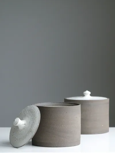 Two grey stoneware lidded jars on a white surface, one with its white speckled lid leaning against the front